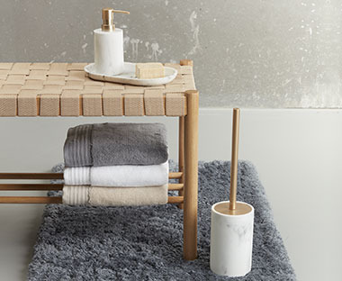 Dispenser di sapone su un vassoio effetto marmo posto su una panca su cui si trava un set di asciugamani, affianco a uno scopino WC su un tappetino per bagno grigior un tapis de bain gris