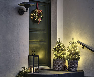Zwei kleine, beleuchtete Kunstbäume und eine Laterne vor einem Hauseingang, an der Tür ein Weihnachtskranz 