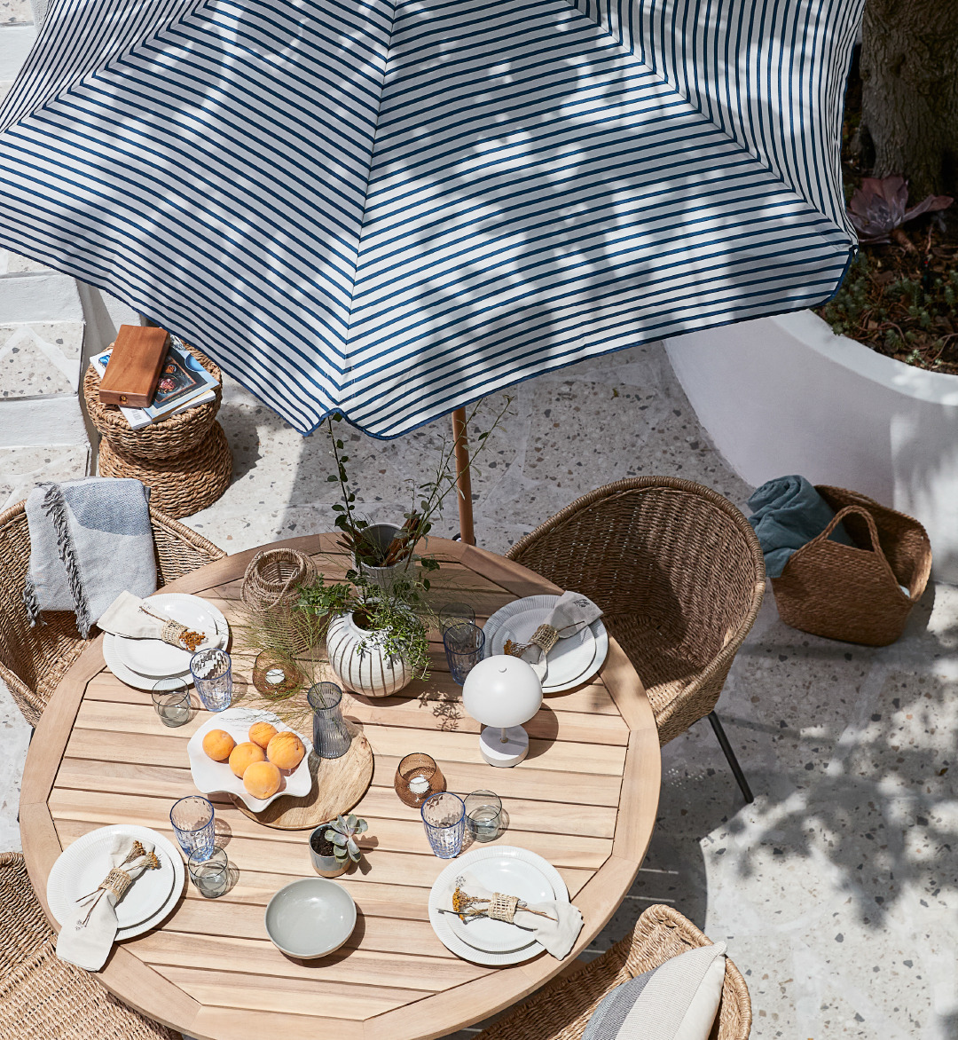 Terrasse avec table en bois bien équipée et chaises en rotin, le tout recouvert d'un parasol.